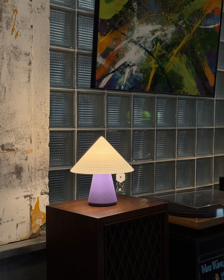 Lavender NOLA lamp on a wood cabinet against a glass-block wall, beside a colorful abstract painting and vinyl records